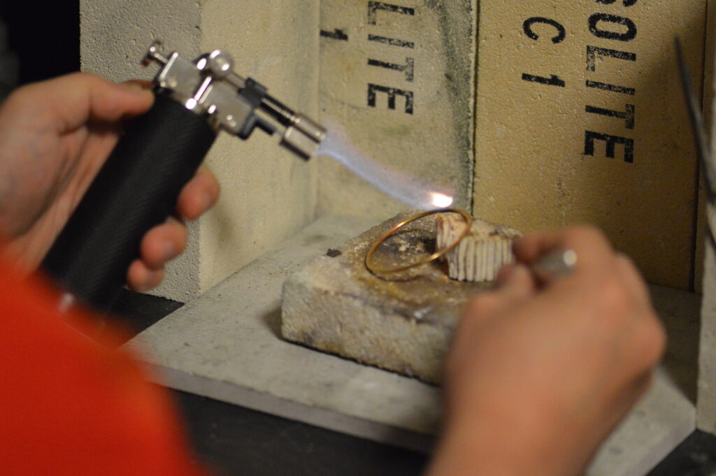 シルバーアクセサリー制作におけるロウ付け工程。バーナーの炎でリングを加熱し、耐火レンガの上で精密に彫金作業を行っている。