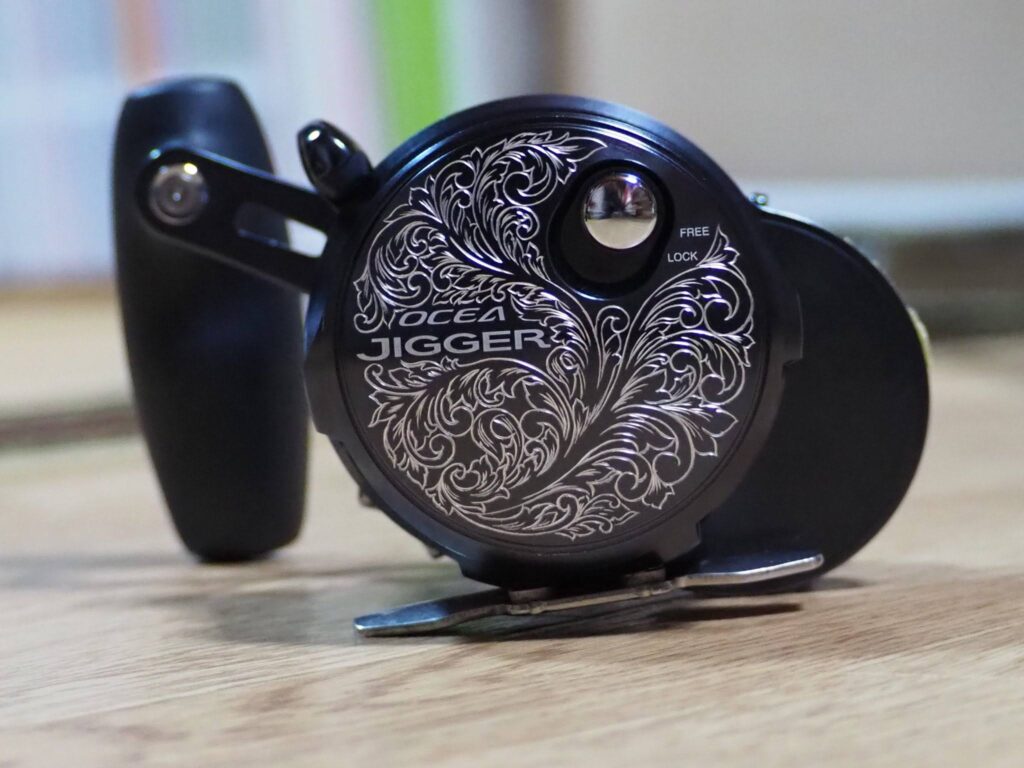 SHIMANO オシアジガーのリール側面に唐草模様を手彫りでエングレービングしたカスタム彫金作品。金属表面に繊細な装飾を施したシルバーエングレービングの事例写真。