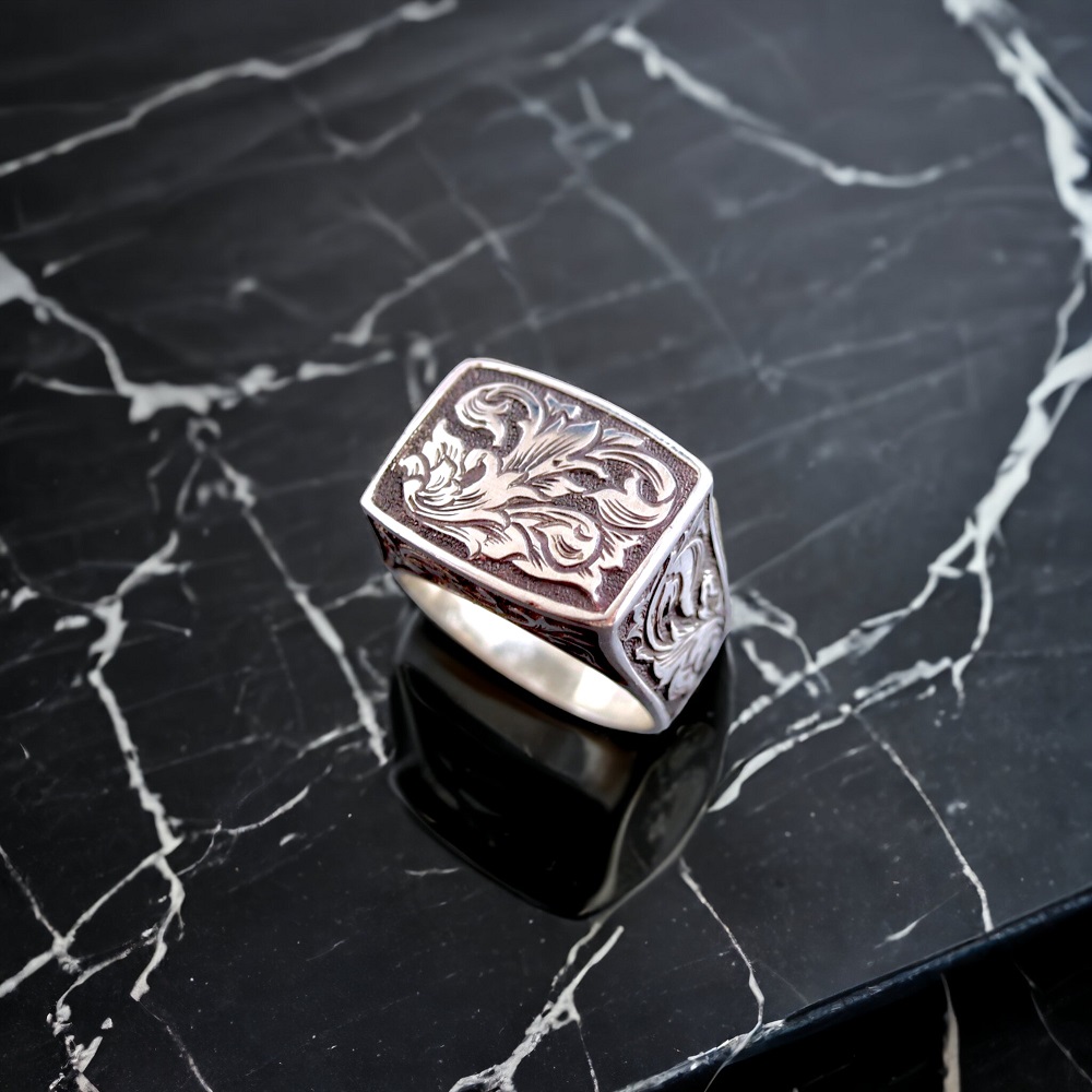 黒い大理石の上に置かれたスクエア型のシルバーリング。手彫りの唐草模様と燻し仕上げによる陰影が際立っている