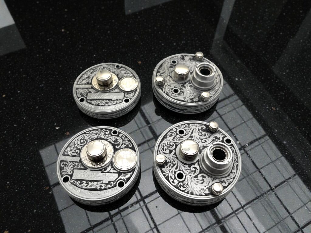 五十鈴工業製ベイトリールのパーツに、唐草文様の手彫り彫金を施した金属部品を4点並べて撮影した写真。黒い光沢面の上に配置されている。