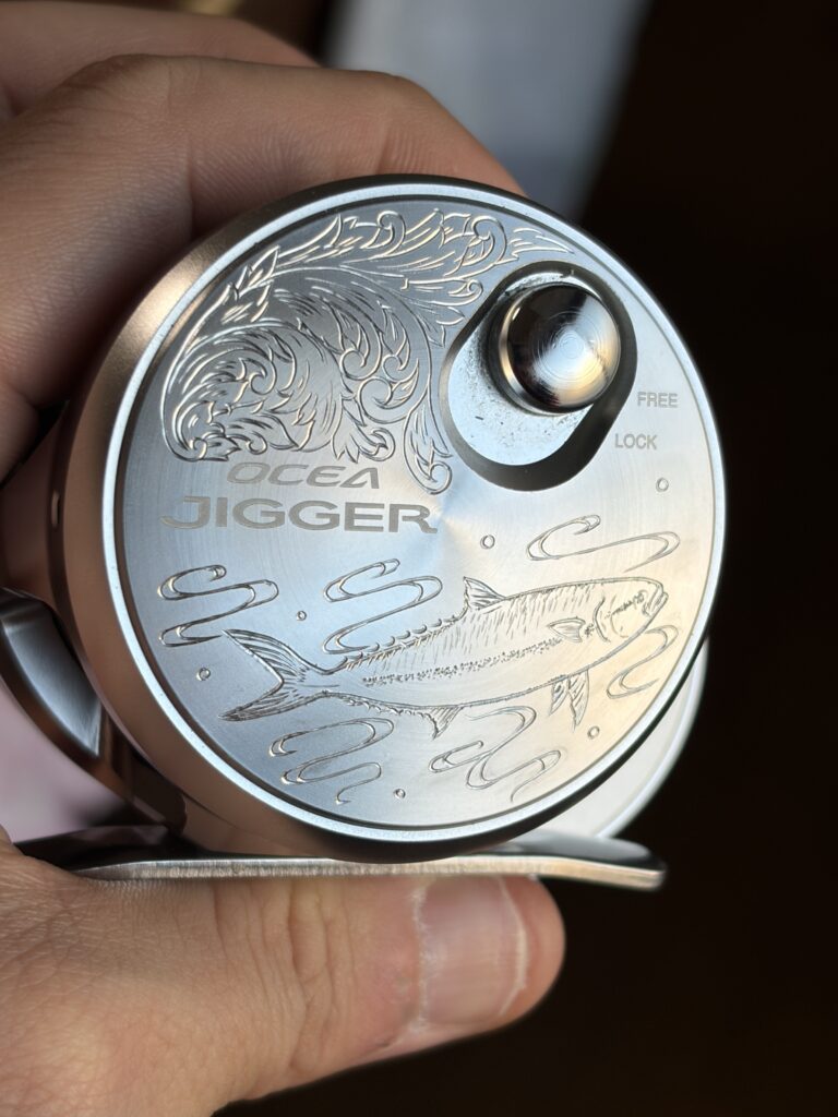 オシアジガーのリール側面カップに、ヒラマサと水流、上部に唐草模様を手彫りで施した金属彫刻のクローズアップ画像